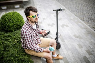 Man with e-scooter relaxing and using smartphone on bench in city. Modern male with electro scooter