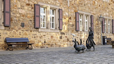 Scene in front of a stone building with shutters, a bench and a statue of a girl with geese, The