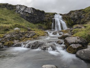 Waterfall cascading over mossy rocks with flowing water and greenery under a cloudy sky. Iceland