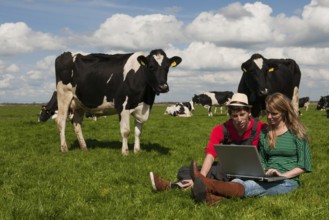 Young couple farmers in farm field with laptop and black and white cows