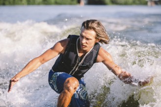 Beautiful long haired blond wakesurf in vest on board along waves of lake. Athletic male athlete