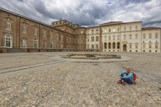 Woman sitting on a pebble square in front of a large building, Palace of Venaria Reale, Residences
