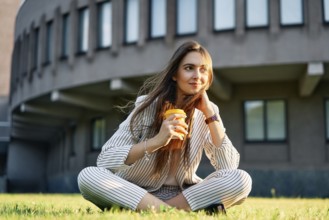 Young stylish woman drinking coffe and relax sitting on a grass in city park. Break time concept