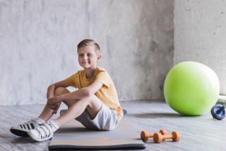 Smiling boy sitting exercise mat with dumbbell pilates ball roller slide