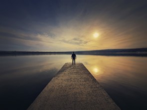 Man standing alone on the pier facing the sunset reflected in the calm lake water. Scenic view with