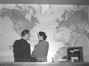In the 1950s. A young couple at a travel agency stands in front of a world map and looks at all the
