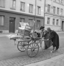 Moving in the 1940s. A couple are pushing a carriage in front of them with some furniture on it.
