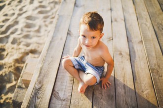 Little Caucasian boy child sitting on wooden pier sandy beach, summer time, sea holiday near water.