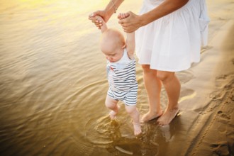 Baby boy walking along beach with mom's support. Mother teaching baby to walk. Outdoor, trust
