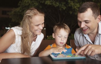 Young parents and little son in a cafe in the evening. Father and son using touch pad, mother