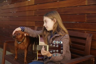 Blond kid girl playing guitar with dog and winter beret on wooden background