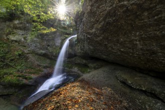 A waterfall flows over mossy rocks, illuminated by sunbeams breaking through the leaves,