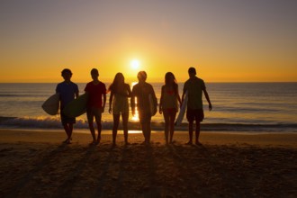 Surfers teen boys and girls group walking on beach at sunshine sunset backlight