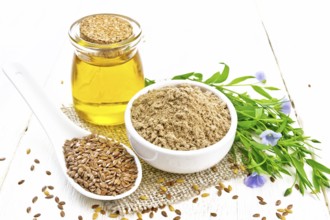 Flaxseed flour in a bowl, linenseeds in a spoon, oil in glass jar on burlap napkin, leaves and blue