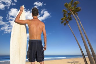 Boy surfer back rear view holding surfboard on California palm trees beach