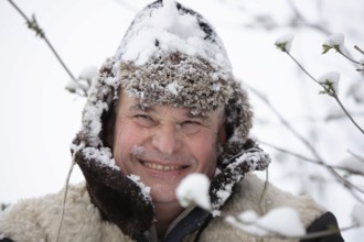 An elderly man in a snowy winter hat smiles and looks into the camera