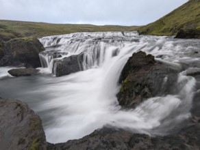 Water cascades over rocks into a flowing river in a serene natural setting. Iceland