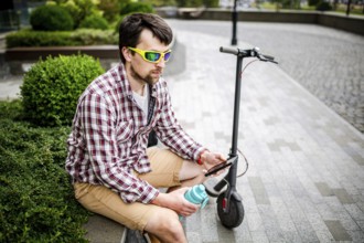 Man with e-scooter relaxing and using smartphone on bench in city. Modern male with electro scooter