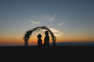 Silhouettes of bride and groom holding hands near the wedding arch at sunset at the wedding