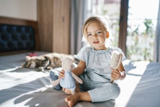 Little smiling girl sitting on the bed with a teddy bear and a comb in her hands.