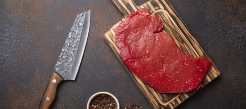 Raw uncooked top round beef steak on wooden cutting board with big kitchen knife and pepper on dark