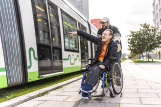 Disabled man in a wheelchair points at something while his friend stands behind him near a tram