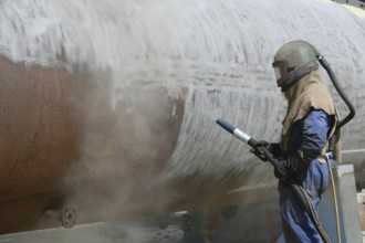 A man weating full safety gear sandblasts the steel casing of a meat drying machine for a rendering