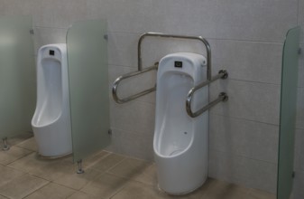 Porcelain urinal with handrails for use by disabled person next to non-disabled urinal in public