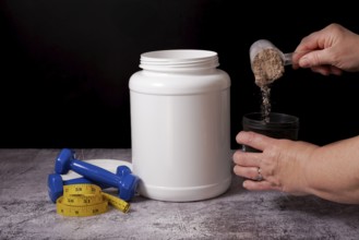 Woman pouring protein powder into a shaker with a spoon, next to a bottle of protein, dumbbells and