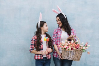 Mother holding tulips basket looking girl holding easter eggs hand against blue background
