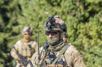 A soldier in camouflage uniform with tactical gear and sunglasses patrols a forest area
