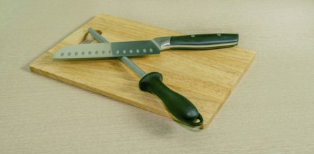 Santoku knife and honing steel on woodblock cutting board with light wood grain background