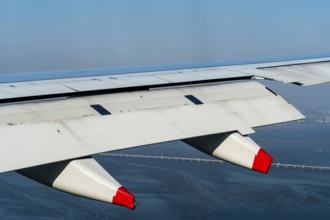 Aircraft wing with red tipped pillion in flight above ocean coastline with city buildings and