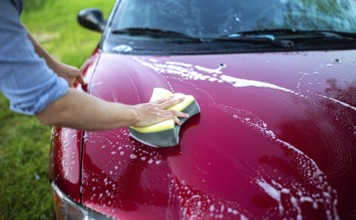 Hand using a sponge to wash the hood of a red car with soap and suds