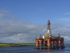 A single oil drilling platform sits in the calm sea with a slightly cloudy landscape behind it,