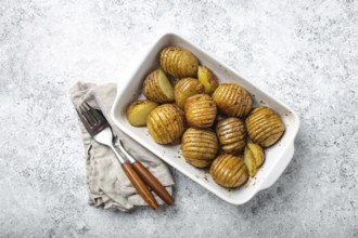 Hasselback potatoes with herbs in white ceramic casserole dish on white concrete table background