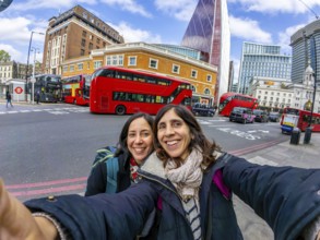 Two happy female tourists taking a selfie with red double decker buses and modern skyscrapers in