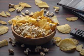 Wooden bowl with crisps and popcorn scattered on the table with remote control and DVDs