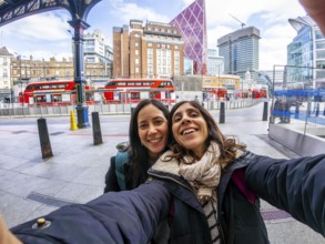 Two happy tourists taking a selfie in the city of london, with red buses and modern buildings in