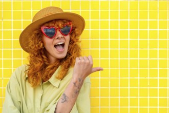 Excited young woman with red hair, heart shaped sunglasses, and straw hat pointing her thumb to the