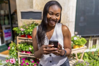 Smiling black business owner using mobile phone app managing online orders in front of her flower