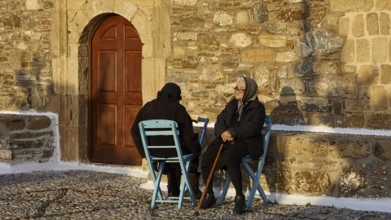 Two elderly people sitting on blue chairs in front of a stone wall and talking, Easter 2025, Church
