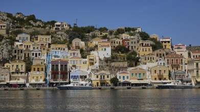 Row of colourful houses along the harbour with boats in the calm water, Symi Town, Symi,