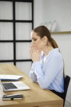 A business woman sits at her desk with a laptop, hands clasped in front of her mouth, visibly deep