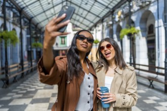 Two women are taking a selfie with a smartphone while walking down the street, holding reusable