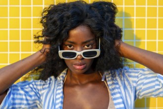 Fashionable model with curly hair touching her head while wearing white sunglasses and posing in
