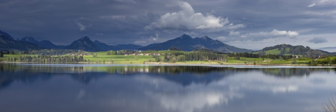 Panorama at sunrise over Lake Hopfensee to the sunlit western shore with farms, behind it receding