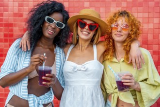 Three multi ethnic young women hugging each other and drinking juice with straw, standing against a