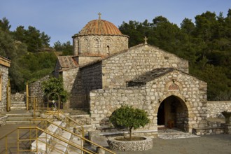 Older Byzantine church with a round tiled roof and a cross on the dome, surrounded by trees and