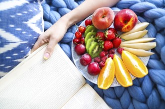 Plate with fresh fruit salad on a blue plaid and book. Woman reading the book and eating fresh